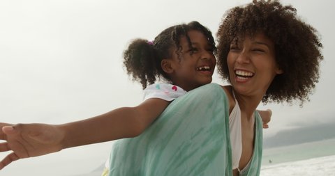 Side view close up of an African American woman in standing by the sea piggybacking her young daughter who has her arms outstretched, both smiling, slow motion