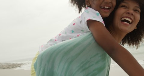 Portrait close up of an African American woman in standing by the sea piggybacking her young daughter and both looking to camera smiling, slow motion