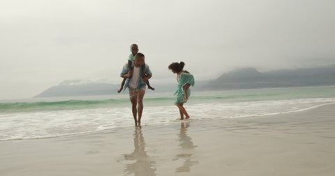 Front view of a smiling African American couple running on a beach away from the sea, piggbacking their young children, dad carrying their son, and mum carrying their daughter, slow motion