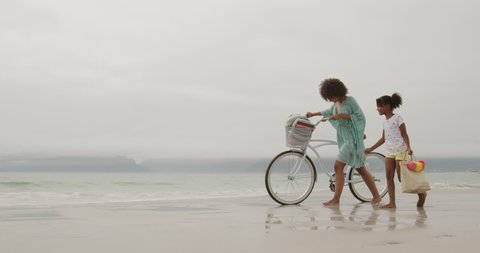 Side view of a smiling African American woman walking with her young daughter and wheeling a white bike along a beach by the sea, slow motion