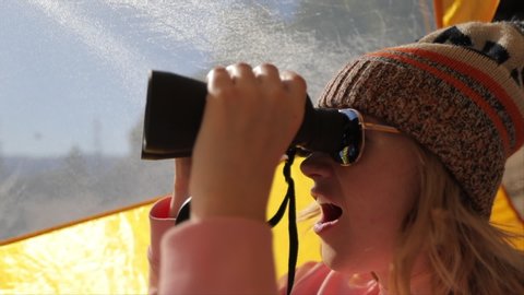 The girl looks through big black binoculars from inside a yellow tourist tent. She puts down the binoculars and in her sunglasses the reflection of Half Dome rock in Yosemite National Park is seen.