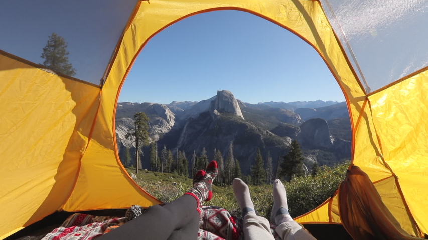 Camping rest in Yosemite Valley. A couple lay inside the yellow tent and point their fingers at the famous Half Dome rock that is seen outside on a picturesque mountain ridge. Yosemite National Park