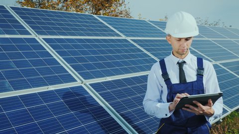 An engineer in a helmet and overalls uses a tablet on the background of a solar power station
