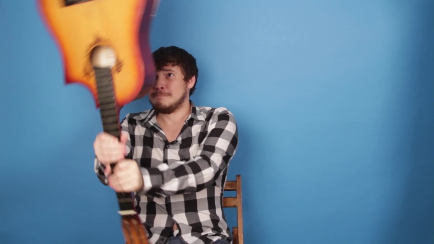A mad man hits himself on the head with a guitar. The guy breaks a guitar on his head on a blue background. Royalty-Free Stock Footage #1042089466