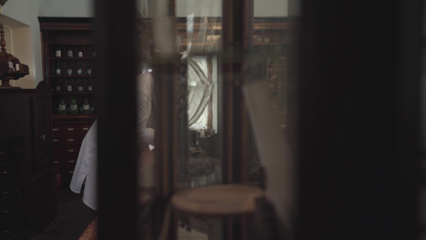 Side view of old energetic Caucasian man in white robe writing with feather pen. Pharmacist staying at cash counter revising his notes. Retro drugstore, vintage pharmacy, pharmaceutical museum.