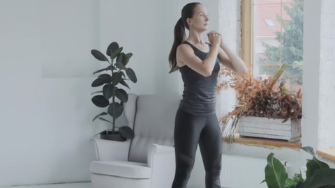 Young fit brunette beautiful girl in gray top, leggings, black sneakers does sport exercises in bright loft style room. Doing sports at home. Healthy lifestyle, training, fitness, weight loss. Squats