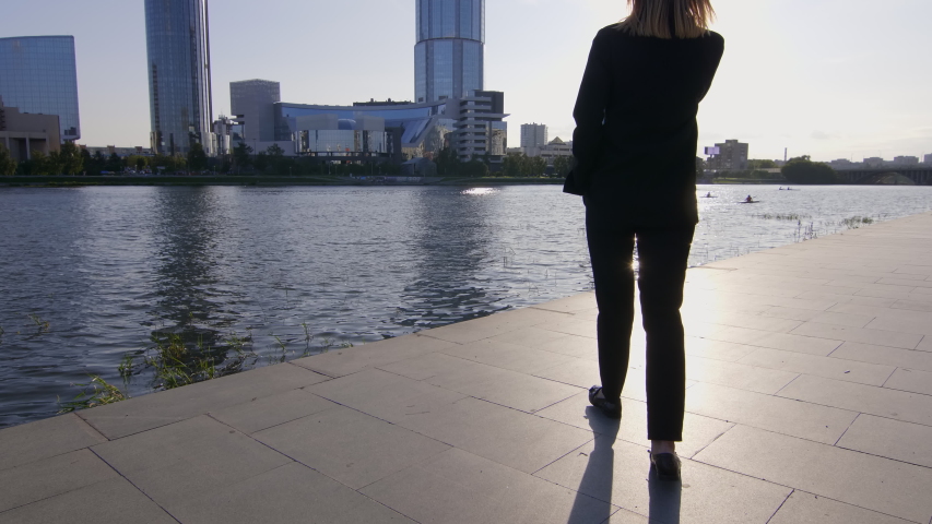 Financial specialist talking by smartphone with her business partner and walking near river. Skyscrapers in background