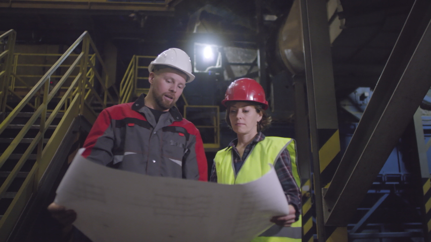 Waist-up arc shot of Caucasian man and woman in hardhats, overalls and high-vis vest standing at industrial facility, holding large paper sheet with technical plan and discussing manufacturing process | Shutterstock HD Video #1042105075