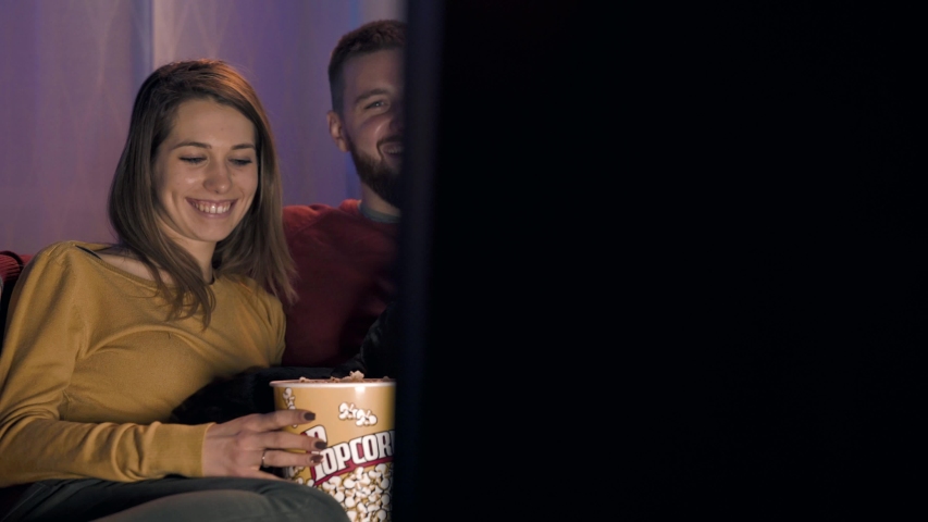 Happy couple at home relaxing on the sofa, they are watching their favorite TV shows and eating popcorn with their dog | Shutterstock HD Video #1042113052