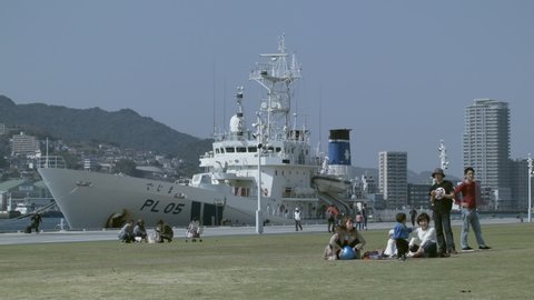 Nagasaki / Japan - 03 24 2019: Group of adults and children on green grass with a docked boat in Nagasaki port in the background, Japan