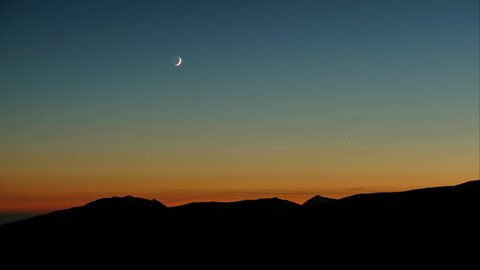 Time lapse of a crescent moon rising over the mountains from dark to morning light