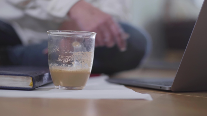 Close-up of coffee cup standing at the foreground. Mature female Caucasian hand closing laptop at the background and taking cappucino to drink. Retiree in casual clothes resting at home.