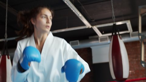 Low angle view of two young female karatekas wearing white kimono and boxing gloves training kicks and punches inside boxing ring