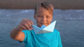 Closeup view of blue paper boat in one small hand of young white boy isolated on sunny early morning sea beach background. Focus at ship, blurry face of smiling kid. Real time 4k video footage.