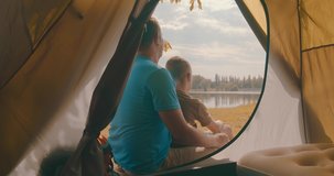 Happy bonding traveler father and son sitting near tent camp around mountains lake under sun light enjoying the leisure and freedom. 4K slow motion video
