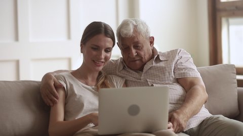 Happy 80s man embracing grown up daughter, sitting together on couch, looking at computer screen. Smiling young woman showing old father how to use applications on laptop, relaxing on sofa at home.