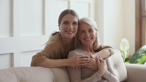 Smiling young woman embracing from back sitting on cozy couch happy affectionate older retired mother. Cheerful two generations family looking at camera, enjoying sweet tender moment together at home.