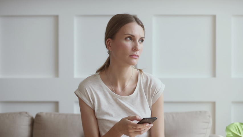 Upset young woman sitting on couch, holding smartphone in hands, received message with bad news. Stressed 30s lady thinking over sudden problems or relations break up after reading sms on cellphone. Royalty-Free Stock Footage #1042130776