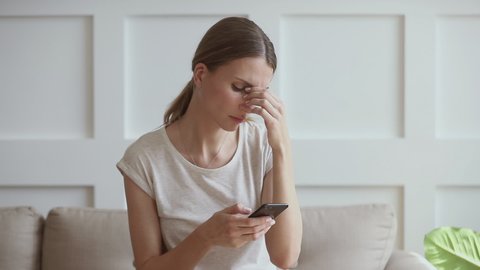 Upset young woman sitting on couch, holding smartphone in hands, received message with bad news. Stressed 30s lady thinking over sudden problems or relations break up after reading sms on cellphone.