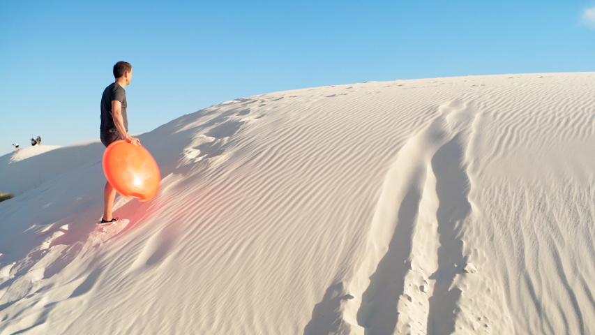 Happy man on sand in white sands dunes national monument park in New Mexico on disk sled sliding down hill panning pov
