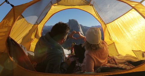 Two people in love meets dawn in the Yosemite Valley. Cosy yellow tent is lit with the rays of the rising sun. The couple make a hand heart with their hands on the background of the landscape. 4K