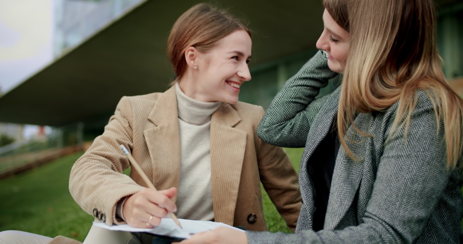 Two pleasure mixed race students female doing homework exercise outside campus on grass, young girls studying together solving mistakes improving knowledges for university exam test, dressed in jacket