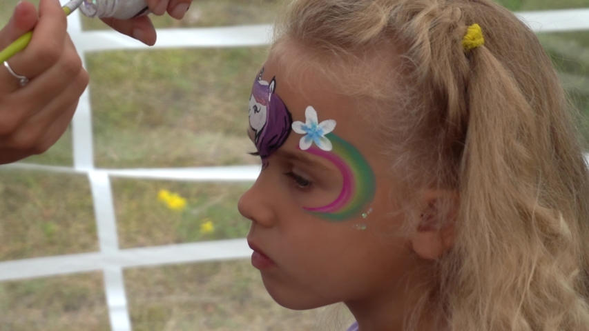 Cute little girl gets a face painting. Hand apply pony with flower and rainbow on child forehead. Camera motion shot with gimbal.