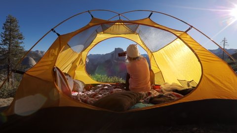 The girl meets morning in a tent in the territory of Yosemite National Park, against the backdrop of the mountain range and the Half Dome cliff, and shoots on a smartphone the landscape around.