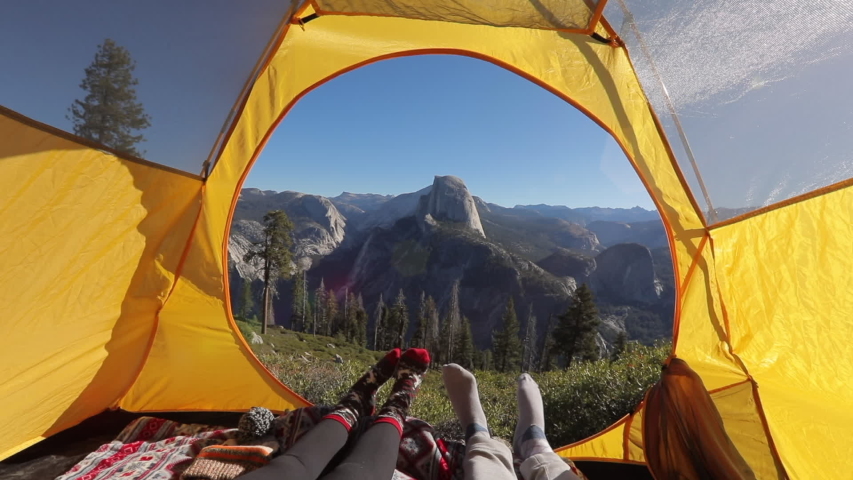 Two pairs of legs in the opening of the tent, against the backdrop of the mountain landscape of Sierra Nevada and the cliff of Half Dome.The sun illuminates the tent.Outside landscape looks inspiring.