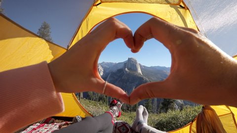 A couple has a day out in Yosemite Valley, admiring the beauty of Sierra Nevada mountains and the Half Dome rock from the tourist tent. They make a hand heart against the backdrop of the landscape.