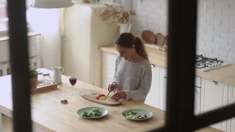 Top view young smiling mixed race woman standing at countertop, chopping vegetables in modern kitchen. Positive millennial lady preparing food alone at home, dreaming about romantic evening.