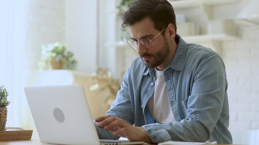 Concentrated young man freelancer in eyeglasses looking at laptop screen, working remotely at home. Head shot focused smiling guy chatting in social network, typing message, internet web surfing. Royalty-Free Stock Footage #1042155763