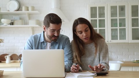Pleasant focused young mixed race family spouses calculating payments, monthly expenses, using computer banking app. Married couple managing budget, counting bills, doing paperwork together at home.