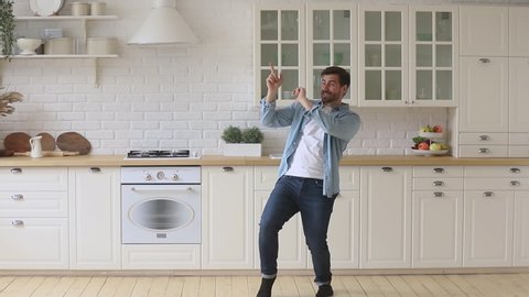 Full length overjoyed young funky guy funny dancing to popular music, celebrating freedom in kitchen. Happy active carefree millennial man having fun alone at home, enjoying free weekend time.