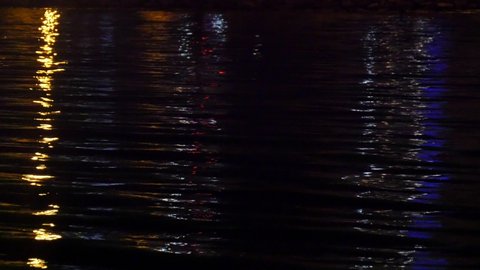 Close-up of reflection of city illumination on sea surface in darkness. Beautiful view of colorful glowing lights reflecting on dark water rippling and sparkling at night.