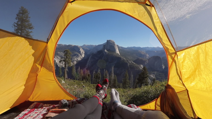 Two pairs of legs in the opening of the tent, against the backdrop of the mountain landscape of Sierra Nevada and the cliff of Half Dome.The sun illuminates the tent.Outside landscape looks inspiring.