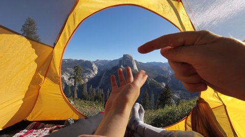 Camping rest in Yosemite Valley. A couple lay inside the yellow tent and point their fingers at the famous Half Dome rock that is seen outside on a picturesque mountain ridge. Yosemite National Park