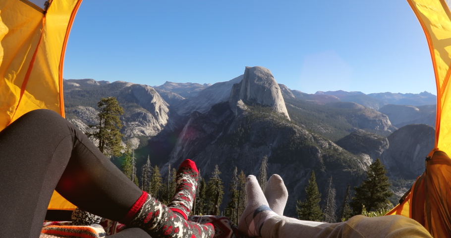 Camping rest in Yosemite Valley. A couple lay inside the yellow tent and point their fingers at the famous Half Dome rock that is seen outside on a picturesque mountain ridge. Yosemite National Park