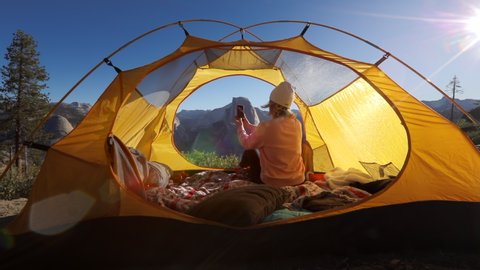 The girl meets morning in a tent in the territory of Yosemite National Park, against the backdrop of the mountain range and the Half Dome cliff, and shoots on a smartphone the landscape around.