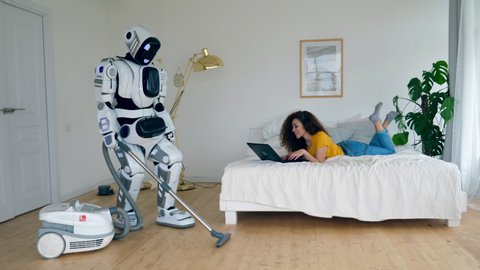 Girl works with a laptop while a cyborg vacuum cleans. Robot and human cooperation.