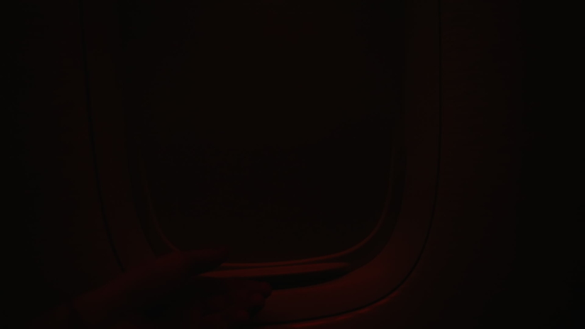 Hand opens airplane window to looks out during air travel. Passenger pov through plane window flight during sunset or sunrise. Close up of porthole of flying airplane at night. Aircraft above clouds. Royalty-Free Stock Footage #1042177981