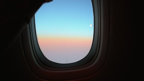 Hand opens airplane window to looks out during air travel. Passenger pov through plane window flight during sunset or sunrise. Close up of porthole of flying airplane at night. Aircraft above clouds.