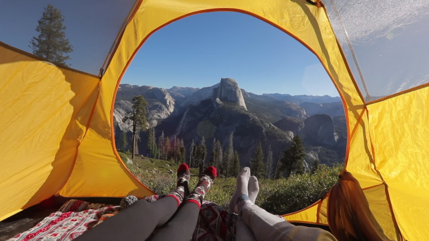 Two pairs of legs in the opening of the tent, against the backdrop of the mountain landscape of Sierra Nevada and the cliff of Half Dome.The sun illuminates the tent.Outside landscape looks inspiring.