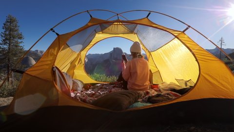 The girl shoots scenic views of the Yosemite Valley, Sierra Nevada ridge and the Half Dome cliff on a smartphone. She sits in a tourist tent flooded with the rays of rising sun. USA.