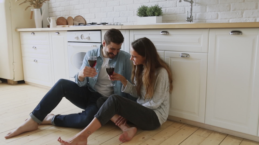 Full length happy young married couple sitting on warm heated floor. clinking glasses with wine, celebrating new house purchase, successful mortgage investment. Laughing family spouse enjoying dating. | Shutterstock HD Video #1042185604
