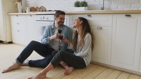 Full length happy young married couple sitting on warm heated floor. clinking glasses with wine, celebrating new house purchase, successful mortgage investment. Laughing family spouse enjoying dating.