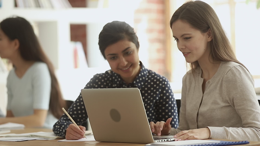 Focused young indian woman working on school project with caucasian friend on computer. Happy diverse girls studying together in library. Mixed race students preparing for exams, writing notes. Royalty-Free Stock Footage #1042185703