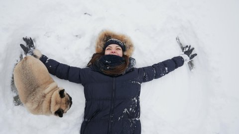 TOP view: Happy woman making snow angels, her pug dog funny running and jumping around