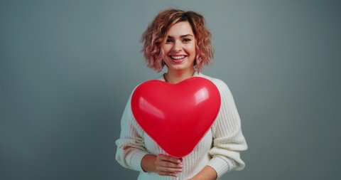 Valentine's Day happy young woman holds red heart balloon smiling and rhythmically moves forward to camera at friend. Symbol of love. Holiday of all lovers. Feelings, emotions. Copy space. Lifestyle