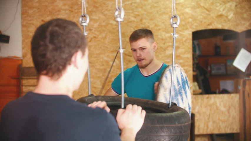 A man knight training on swords on the bunch of tires in the gym - another man holding the bundle